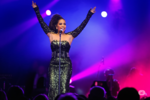 Alcione Show Curitiba Inédito: Alcione, a renomada cantora brasileira, encanta o público durante sua apresentação inédita em Curitiba, em um palco vibrante com iluminação colorida.